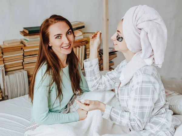 Yüzün ve saçın güzelliği için iki kız maske ve yama yapıyor. Kadınlar genç cildine özen gösterirler. Kız arkadaşlar gülüyor, surat asıyor ve kafalarında havlularla evde güzellik tedavisi yapıyorlar..