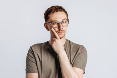 Gözlüklü yakışıklı genç bir adam bir karar vermeyi düşünüyor ve elini yüzüne dayıyor. Kahverengi gömlekli, gri arka planda poz veren esmer iş adamı. Reklam için yer.