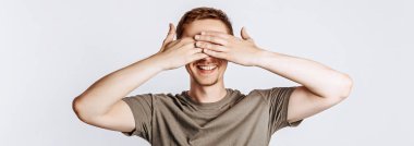 Yakışıklı genç adam gözlerini iki eliyle kapar ve gülümser. Yeşil gömlekli, gri arka planda poz veren esmer bir kadın. Reklam için yer var. Göremiyorum..