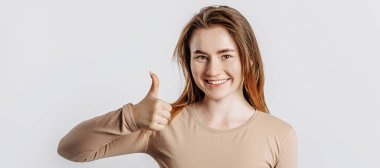 Güzel genç kız gülümsüyor ve beyaz izole bir arkaplanda baş parmak işareti yapıyor.
