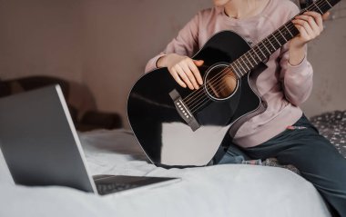 Evde online öğrenmenin yardımıyla gitar çalmayı öğrenen bir kız. Yatakta dizüstü bilgisayarı ve siyah gitarıyla oturan bir kadın.