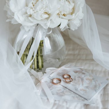 Bir vazo içindeki şakayık çiçeği buketi yeni evlilerin yatağında duruyor. Gelinin peçesinin arkasında da davetiyeler ve yüzükler var.