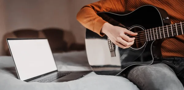 Man learning to play guitar with the help of online learning at home. Guy sitting on bed with laptop and black guitar