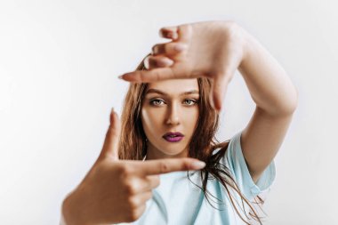 Güzel genç kız, elleri izole edilmiş gri bir arka planda bir çerçeve yapıyor. Mor dudaklı, moda makyajlı kadın kameraya ciddi ciddi bakıyor. Reklam için yer.