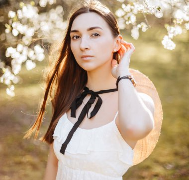 Baharda parkta hasır şapkalı bir kız. Uzun saçlı esmer kadın, yaz doğasının arka planında bir şapka tutuyor. Gençlik ve güzellik.