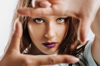 Güzel genç kız, elleri izole edilmiş gri bir arka planda bir çerçeve yapıyor. Mor dudaklı, moda makyajlı kadın kameraya ciddi ciddi bakıyor. Reklam için yer.