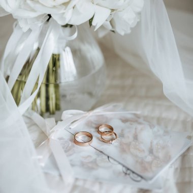 Bir vazo içindeki şakayık çiçeği buketi yeni evlilerin yatağında duruyor. Gelinin peçesinin arkasında da davetiyeler ve yüzükler var.