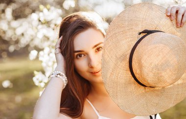 Baharda parkta hasır şapkalı bir kız. Uzun saçlı esmer kadın, yaz doğasının arka planında bir şapka tutuyor. Gençlik ve güzellik.