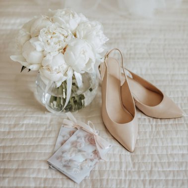 Düğün buketi şakayık çiçekleri vazoda duruyor. Yeni evlilerin yatağında duruyor. Gelinin arka planında da davetiyeler ve ayakkabılar var.