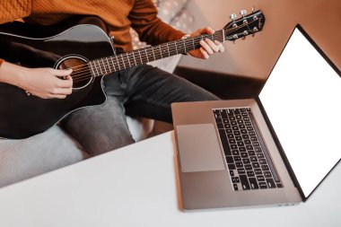 Evde online öğrenmenin yardımıyla gitar çalmayı öğrenen bir adam. Laptop ve siyah gitarlı bir adam masada oturuyor.