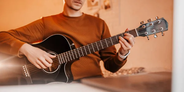 Man learning to play guitar with the help of online learning at home. Guy sitting at table with laptop and black guitar