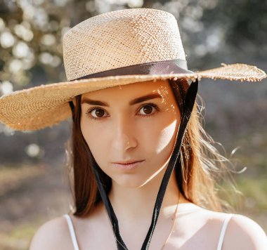 Baharda parkta hasır şapkalı bir kız. Kahverengi gözlü bir esmer yazın doğanın arka planında kameraya bakar. Gençlik ve güzellik.