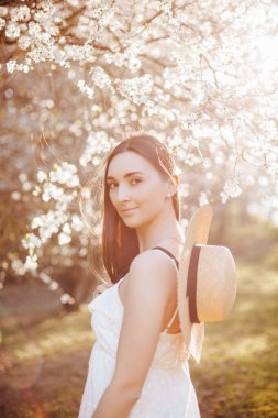 Baharda parkta hasır şapkalı bir kız. Uzun saçlı esmer kadın, yaz doğasının arka planında bir şapka tutuyor. Gençlik ve güzellik.