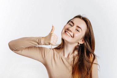 Güzel genç esmer kız gülümsüyor ve beyaz izole bir arka planda baş parmak işareti yapıyor.