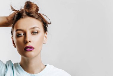 Güzel esmer bir kız saçını tutup kendi saç stilini yapar. Uzun saçlı genç bir manken, gri arka planda mor dudaklı bir moda makyajı. Kadın cidden kameraya bakıyor..