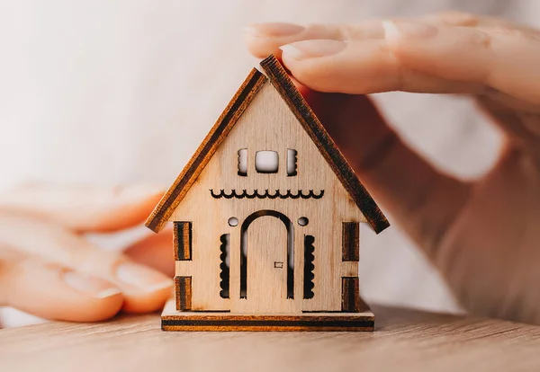 Woman holds and protects a wooden house with her hands with the sun on a light pink background. Sweet home