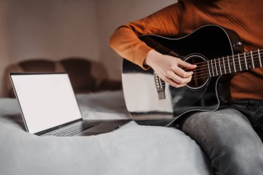 Evde online öğrenmenin yardımıyla gitar çalmayı öğrenen bir adam. Laptop ve siyah gitarla yatakta oturan adam.