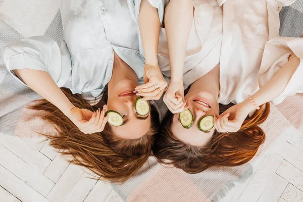 两个女孩做了自制的脸和头发美容面具 黄瓜的新鲜的皮肤周围的眼睛 女人照顾年轻的皮肤 女朋友们在家里躺在地板上躺在枕头上笑 — 图库照片