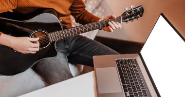Evde online öğrenmenin yardımıyla gitar çalmayı öğrenen bir adam. Laptop ve siyah gitarlı bir adam masada oturuyor.