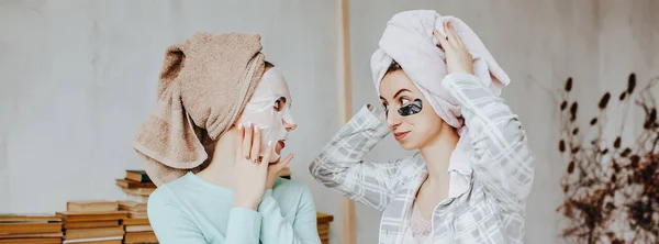 Yüzün ve saçın güzelliği için iki kız maske ve yama yapıyor. Kadınlar genç cildine özen gösterirler. Kız arkadaşlar gülüyor, surat asıyor ve kafalarında havlularla evde güzellik tedavisi yapıyorlar..