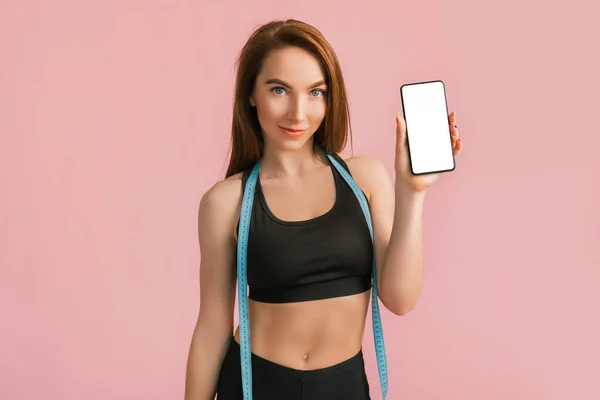 健康女孩笑着拿起电话 拿着一个模型 摆出一副粉红背景的黑色运动服的尺码 身材苗条 体形漂亮 皮肤晒黑的女人 — 图库照片