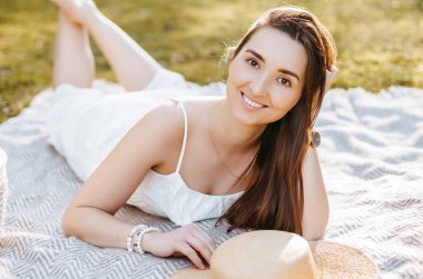 Baharda parkta hasır şapkalı bir kız. Esmer, uzun saçlı, yaz doğasının arka planında bir ekose üzerinde yatıyor. Gençlik ve güzellik.