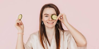 Kız, ev yapımı güzellik maskeleri yapıyor. Salatalık gözün etrafındaki derinin tazeliği için. Kadın genç cildine dikkat eder. Model gülüyor ve pembe arka planda kaplıcada eğleniyor.