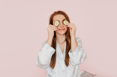 Kız, ev yapımı güzellik maskeleri yapıyor. Salatalık gözün etrafındaki derinin tazeliği için. Kadın genç cildine dikkat eder. Model gülüyor ve pembe arka planda kaplıcada eğleniyor.