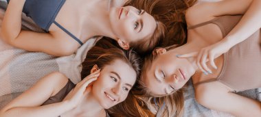 Women rest and have fun. Girlfriends laugh at home lying on the floor on pillows. Tree girls make homemade face and hair beauty masks. Women take care of youthful skin. clipart