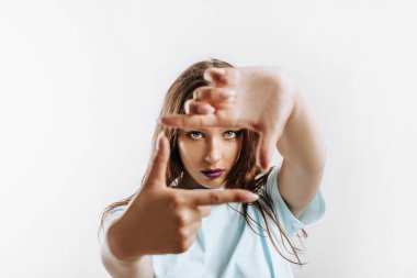 Güzel genç kız, elleri izole edilmiş gri bir arka planda bir çerçeve yapıyor. Mor dudaklı, moda makyajlı kadın kameraya ciddi ciddi bakıyor. Reklam için yer.