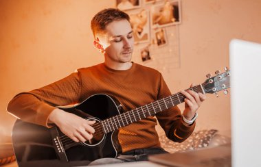 Evde online öğrenmenin yardımıyla gitar çalmayı öğrenen bir adam. Laptop ve siyah gitarlı bir adam masada oturuyor.