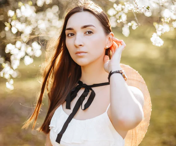 Baharda parkta hasır şapkalı bir kız. Uzun saçlı esmer kadın, yaz doğasının arka planında bir şapka tutuyor. Gençlik ve güzellik.