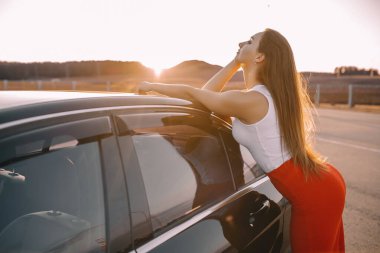 Güzel bir genç kız akşamleyin bir arabanın yanında güneşin batışında boş bir otoparkta.