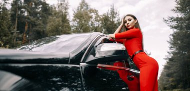 Kırmızı tulumlu güzel bir genç kız ormanda boş bir yolda siyah bir arabanın yanında duruyor.