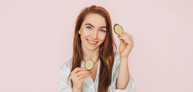 Kız, ev yapımı güzellik maskeleri yapıyor. Salatalık gözün etrafındaki derinin tazeliği için. Kadın genç cildine dikkat eder. Model gülüyor ve pembe arka planda kaplıcada eğleniyor.