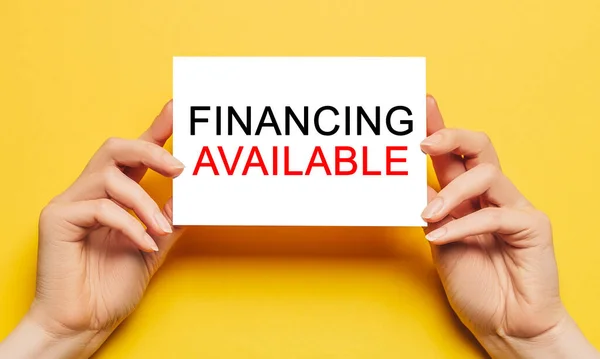 Female hands hold card paper with text Financing Available on a yellow background. Business and finance concept