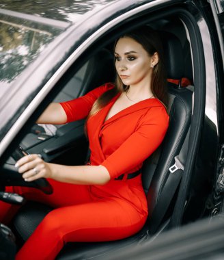 Kırmızı tulumlu güzel bir genç kız ormanda boş bir yolda siyah bir arabanın direksiyonunda oturuyor.