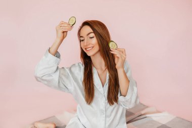 Kız, ev yapımı güzellik maskeleri yapıyor. Salatalık gözün etrafındaki derinin tazeliği için. Kadın genç cildine dikkat eder. Model gülüyor ve pembe arka planda kaplıcada eğleniyor.