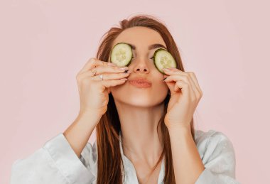 Kız, ev yapımı güzellik maskeleri yapıyor. Salatalık gözün etrafındaki derinin tazeliği için. Kadın genç cildine dikkat eder. Model gülüyor ve pembe arka planda kaplıcada eğleniyor.