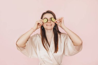 Kız, ev yapımı güzellik maskeleri yapıyor. Salatalık gözün etrafındaki derinin tazeliği için. Kadın genç cildine dikkat eder. Model gülüyor ve pembe arka planda kaplıcada eğleniyor.