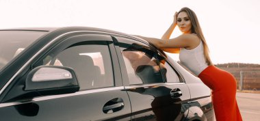 Güzel bir genç kız akşamleyin bir arabanın yanında güneşin batışında boş bir otoparkta.