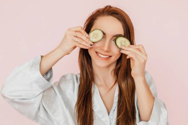 Kız, ev yapımı güzellik maskeleri yapıyor. Salatalık gözün etrafındaki derinin tazeliği için. Kadın genç cildine dikkat eder. Model gülüyor ve pembe arka planda kaplıcada eğleniyor.