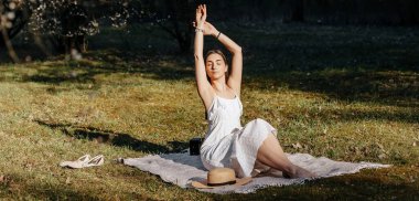 Baharda parkta hasır şapkalı bir kız. Uzun saçlı esmer, yaz doğasının arka planında bir ekose üzerinde oturuyor. Gençlik ve güzellik.