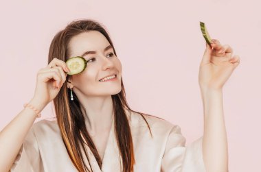 Kız, ev yapımı güzellik maskeleri yapıyor. Salatalık gözün etrafındaki derinin tazeliği için. Kadın genç cildine dikkat eder. Model gülüyor ve pembe arka planda kaplıcada eğleniyor.