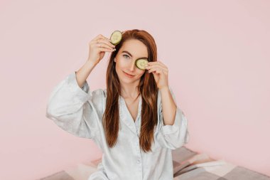 Kız, ev yapımı güzellik maskeleri yapıyor. Salatalık gözün etrafındaki derinin tazeliği için. Kadın genç cildine dikkat eder. Model gülüyor ve pembe arka planda kaplıcada eğleniyor.