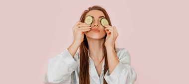 Kız, ev yapımı güzellik maskeleri yapıyor. Salatalık gözün etrafındaki derinin tazeliği için. Kadın genç cildine dikkat eder. Model gülüyor ve pembe arka planda kaplıcada eğleniyor.