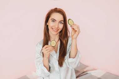 Kız, ev yapımı güzellik maskeleri yapıyor. Salatalık gözün etrafındaki derinin tazeliği için. Kadın genç cildine dikkat eder. Model gülüyor ve pembe arka planda kaplıcada eğleniyor.