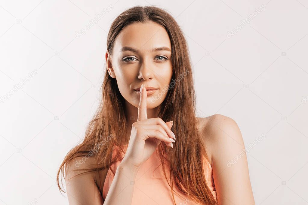 Shh gesture. Young beautiful serious girl holding a finger to her lips on a white isolated background. A woman asks to be silent, a place for advertising. Positive brunette in orange top