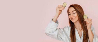 Kız, ev yapımı güzellik maskeleri yapıyor. Salatalık gözün etrafındaki derinin tazeliği için. Kadın genç cildine dikkat eder. Model gülüyor ve pembe arka planda kaplıcada eğleniyor.