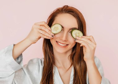 Kız, ev yapımı güzellik maskeleri yapıyor. Salatalık gözün etrafındaki derinin tazeliği için. Kadın genç cildine dikkat eder. Model gülüyor ve pembe arka planda kaplıcada eğleniyor.